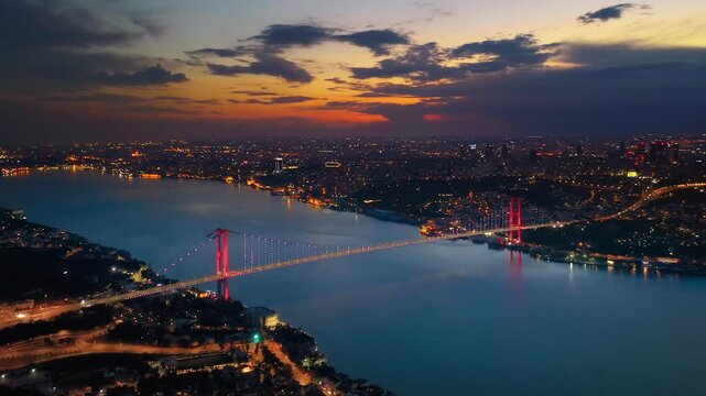 Aerial view of illuminated Bosphorus Bridge spanning Bosphorus at dusk with LED lit red towers city lights and dramatic orange sunset sky over Istanbul