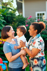 Vertical shot of Caucasian mother holding toddler while African American woman greeting child outdoors, showing family bonding and caregiving. Ideal for parenting, childcare, adoption campaigns