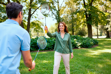 Caucasian middle aged woman and man playing badminton in park, swinging rackets during outdoor recreation. Supporting active lifestyle, wellness, leisure campaign, wide shot