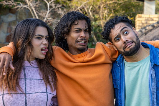 Diverse friends wearing pink sweater, denim shirt, making exaggerated faces in yard near stone wall