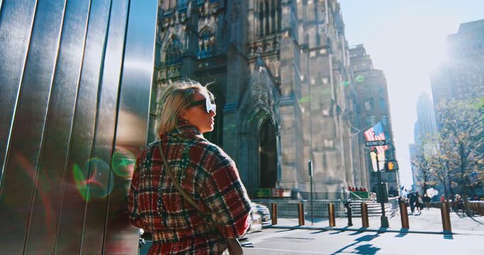 Rear view of a woman in checkered coat leaning on the wall. Lady in sunglasses looks at the cathedral in the backdrop. Low angle perspective.