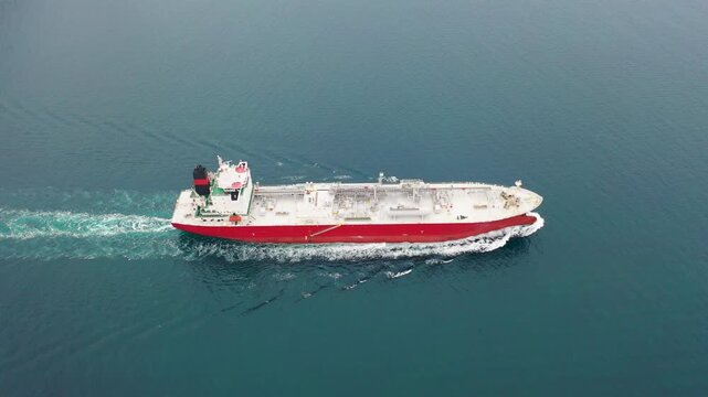 LNG tanker with red hull sailing across open sea in aerial top-down view with white foam wake and complex pipe manifold deck