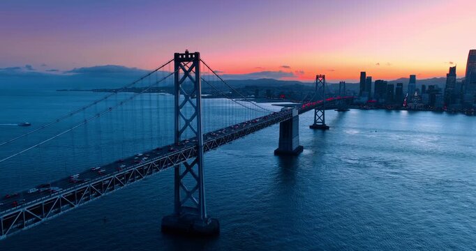 The Oakland Bay Bridge roads are filled with numerous moving cars to and from the downtown. Silhouette of city skyline against pink sky in the backdrop. San Francisco, California, USA at sunset.
