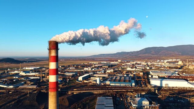 Industrial smokestack emitting white smoke over factory during sunset. Aerial view of striped chimney releasing thick smoke over industrial plant with mountains.