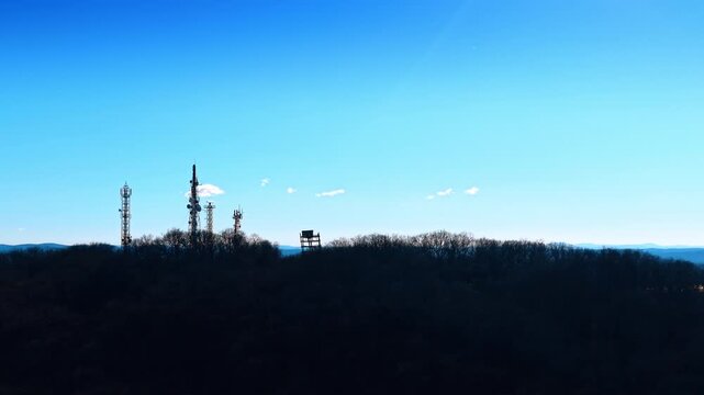 Silhouette of telecommunication masts on hill during sunset. Dark outline of signal towers and antennas on a wooded ridge against a clear sky.