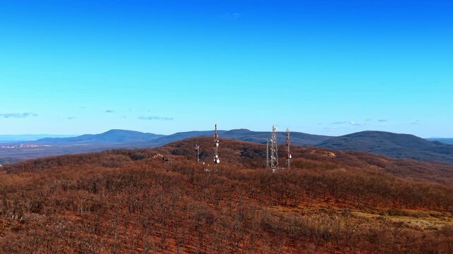 Wide aerial shot of telecommunication towers on wooded ridge. Multiple signal masts with antennas standing on top of a forest hill under blue sky.