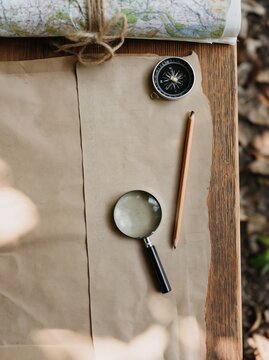 Overhead flat lay of scavenger hunt supplies including a compass, magnifying glass, pencil, and map on a wooden surface with natural elements in the background