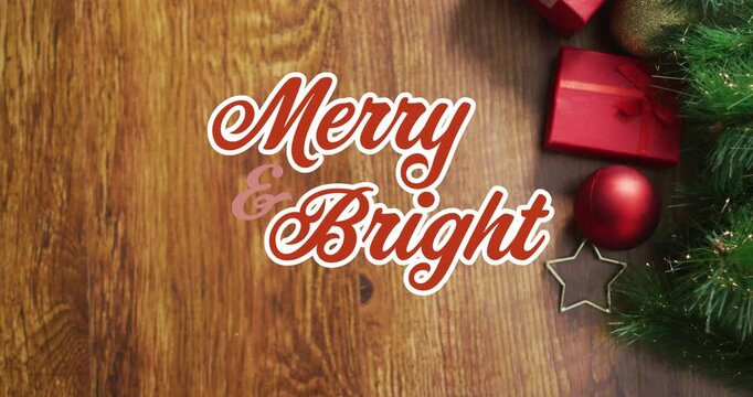 Animation of merry and bright text over christmas tree, gift boxes and bauble on wooden table
