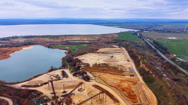 Wide aerial shot of industrial sand quarry near large lake. Panoramic view of open pit mine with heavy machinery, conveyor belts, artificial pond and surrounding fields.