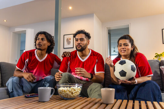 Asian friends sitting on couch in living room wearing red jerseys holding soccer ball with popcorn