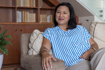 Fototapeta premium Mature woman sitting relaxed on beige sofa in lounge, showing bangle and wristwatch with bookshelf