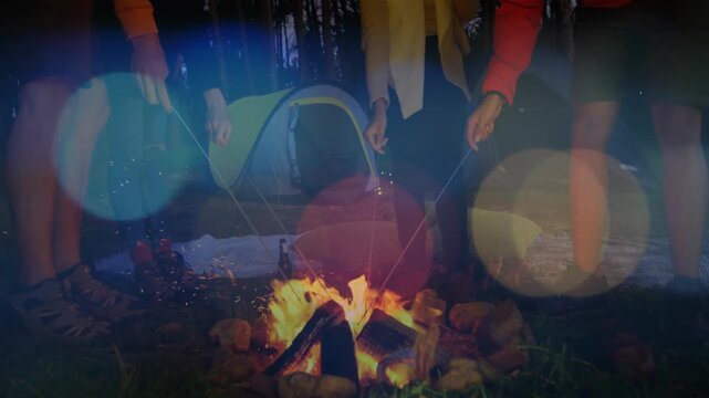 Campers standing holding sticks over fire roasting, flames flaring, embers drifting, bokeh floating