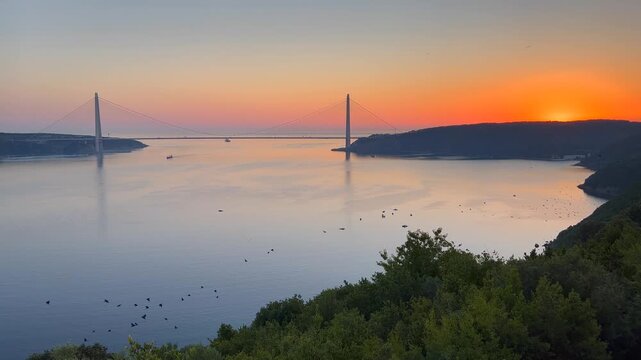 Yavuz Sultan Selim Bridge silhouetted against vibrant orange sunrise over Bosphorus with mirror calm water bird flock and forested hills, Istanbul