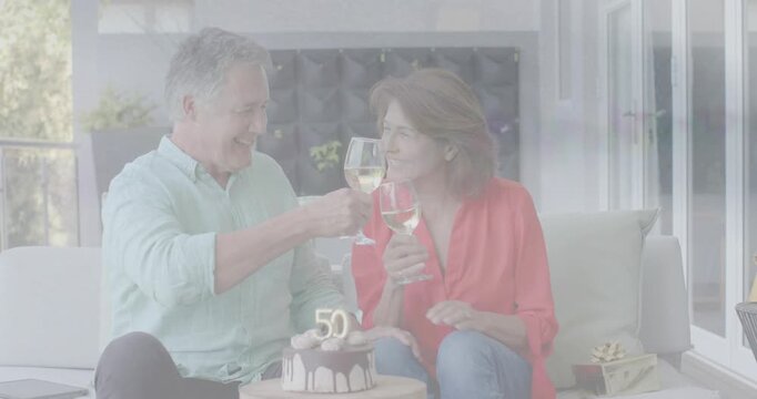 Woman laughing and raising wine glass, man mirroring toast, couple marking 50th anniversary by cake