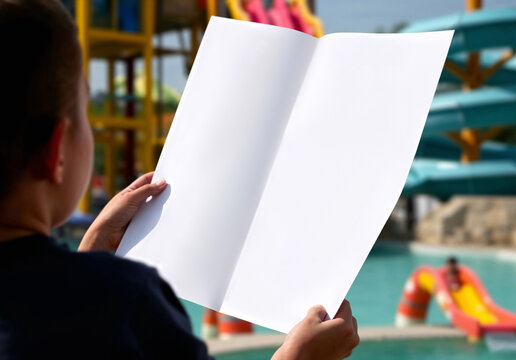 Kid hands holding blank white map mockup, ai generated. Unfolded brochure for swimming waterslide locate, blurred aquapark background. Child lost in resort and planning on vintage list template.