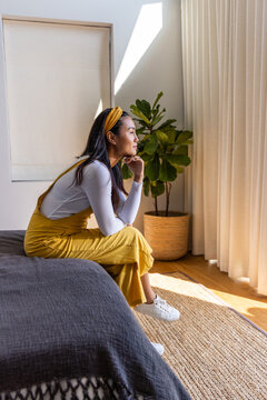 Asian woman 20s sitting on bed edge in bedroom with dark gray blanket wearing mustard pinafore