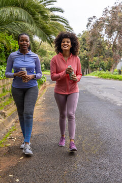 African American female friends in athletic wear walking on suburban road holding water bottles