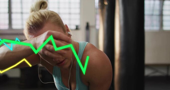 Vertical video: Gasping woman checking metrics in gym after set, wearing tank top and wired earbuds