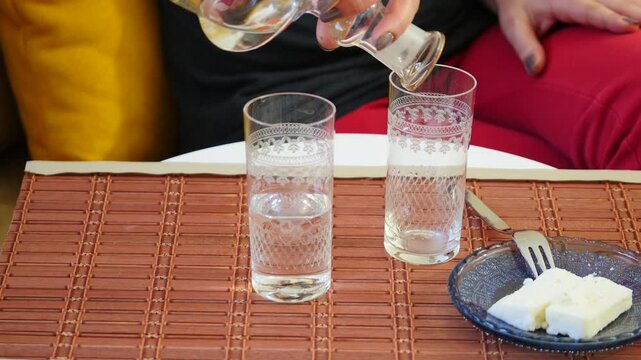 Woman's hand pouring raki from floral glass carafe into traditional engraved glasses with water glass and white cheese on bamboo table