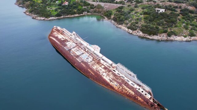 Aerial view of a large, rusty shipwreck lying on its side in a tranquil bay in greece