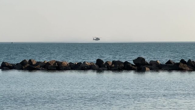 Italian Navy helicopter flying low over the sea beyond a rocky breakwater in the Gulf of La Spezia, Liguria, Italy.