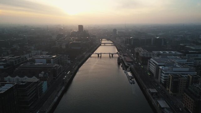 aerial view flying over Liffey River in the heart of Dublin City centre featuring different bridges and a golden dusty sunset, - Dublin, Ireland