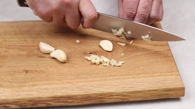 Close up of hand mincing garlic with knife on wooden cutting board during food preparation