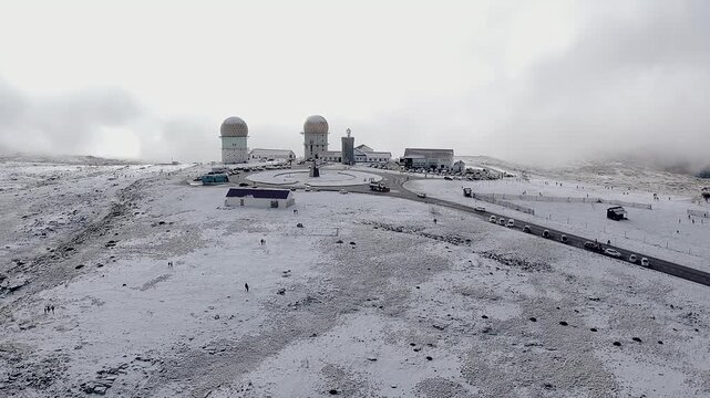 Drone view of Torre, highest point of Serra da Estrela, Portugal, at N339 road, showing radar domes, summit buildings, parked vehicles and snow-covered plateau, aerial orbit
