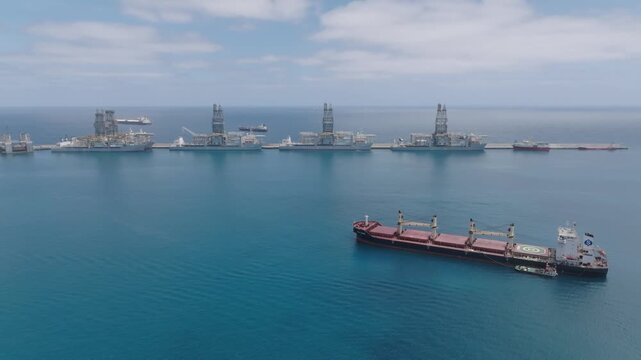 Slow aerial flight from coast toward the open ocean at Las Palmas port, Gran Canaria, featuring Reina Sofia pier with offshore platforms and cargo ship in the Atlantic.