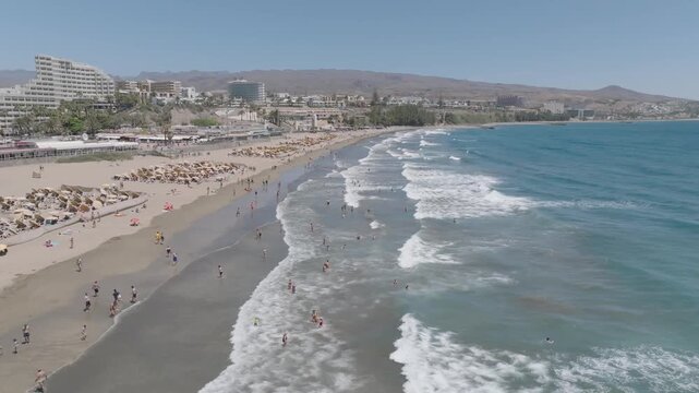 Drone forward flight along Playa del Ingles beach in Gran Canaria, Spain, showing waves, people, sandy shore, and coastal resorts.