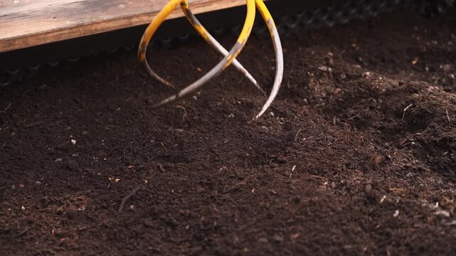 Close up slow motion footage of garden claw tool cultivating and loosening rich soil in raised bed. Backyard gardening, soil aeration and preparation for planting crops.