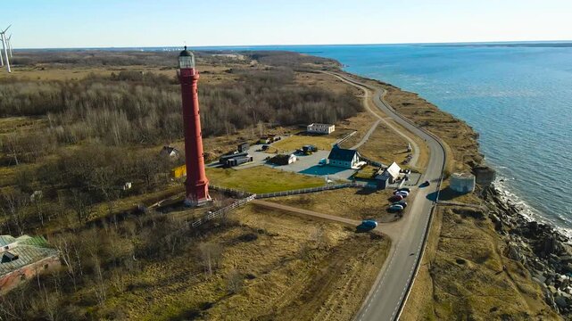 Scenic aerial footage showing the historic red Pakri lighthouse standing on a high limestone cliff coast, with a vast wind farm visible on the horizon during a sunny spring day in Estonia