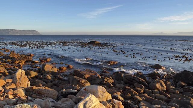 Expansive view of a rugged shoreline of the Cape Peninsula with weathered rocks and the kelpy sea meeting distant hazy mountains under a bright blue sky with wispy clouds.