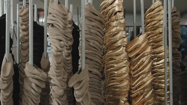 Rows of beige and gold satin ballet pointe shoes stacked and hanging on white metal poles in a dance shoe manufacturing factory