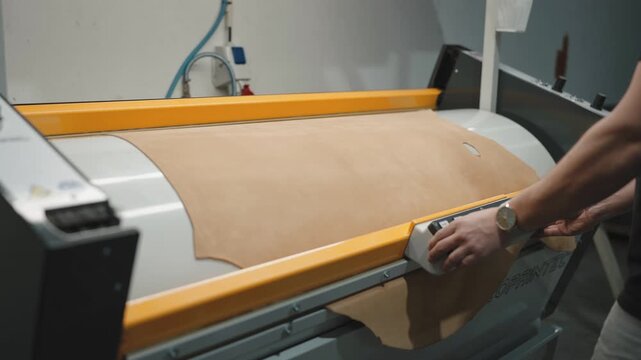 Worker feeds large flat leather hide through yellow industrial press or skiving machine