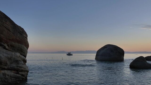 A tranquil scene unfolds as a small boat navigates calm waters near imposing rock formations against a backdrop of mountains in the distance, during a soft sunrise at Millers Point, Simon's Town.