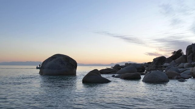 Fishing boat motors homeward through a serene coastal dawn, where smooth boulders rise from the glassy water and a soft golden light spills across the rocky shoreline at Millers Point, Simon's Town.