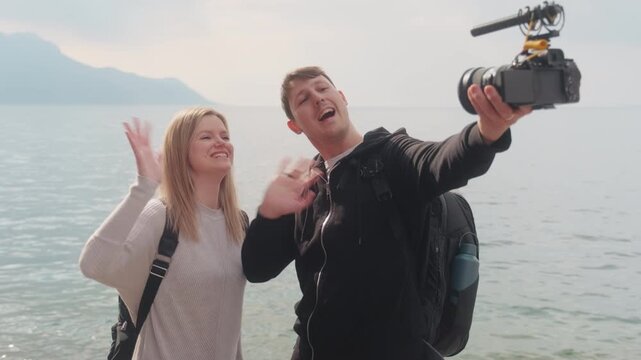 Caucasian couple vlogging at lakeside during the day with mountains in the back, gimbal shot