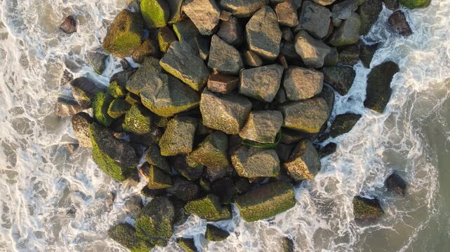 Top-down drone shot of ocean waves crashing against a rocky breakwater on the coast of Sri Lanka, featuring white sea foam and green mossy stones.