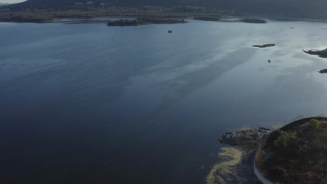 Drone glides forward over Lake Pichola's calm waters dotted with islands, revealing distant Aravalli Range hills under soft sunrise skies with warm golden and pink tones in Udaipur, Rajasthan.