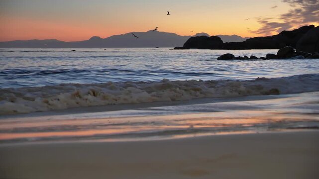 Golden hour light paints the sky with warm hues as gentle waves roll onto a sandy beach, with silhouetted mountains and soaring birds in the background at Millers Point, Simon's Town.