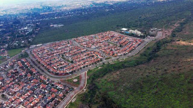 Aerial view of dense suburban housing estate with uniform terracotta rooftops surrounded by green forest and dry hillside on the urban fringe