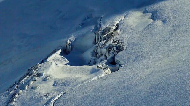 An aerial close-up of a deep glacial crevasse and ice fracture in the Chamonix Alps of France, showing the textured blue ice and snowy terrain of the Mont Blanc massif on a sunny winter day