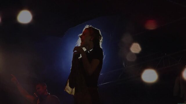 Music cue sending blonde lead flipping hair, singing into mic, backlights forming halo for crowd