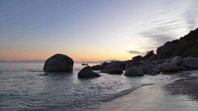 A tranquil beach scene unfolds with a boat passing smooth water-worn boulders scattered along the shore as the sun slowly rises, casting a warm glow over the horizon at Millers Point, Simon's Town.