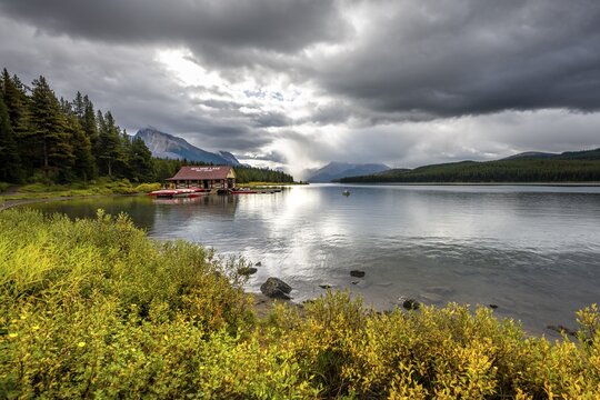 Curly Phillips Boathouse, historic boathouse on the shore of Maligne Lake, behind it mountain range Queen Elizabeth Ranges, autumn vegetation, cloudy sky, Jasper National Park, Rocky Mountains, Alberta, Canada