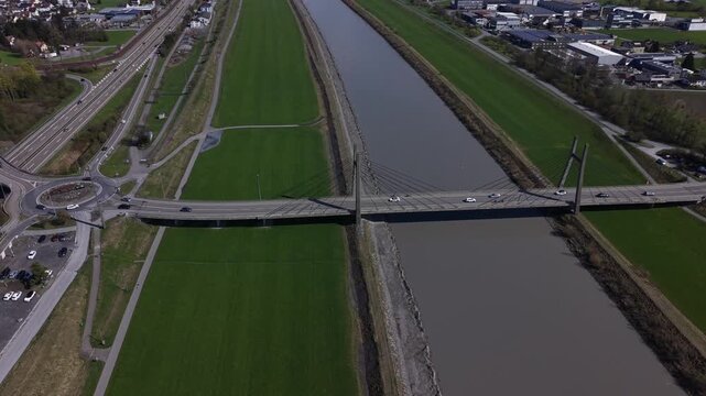 Drone lateral movement capturing complex transport infrastructure including highway, bridge, roundabout, cycling paths, and river crossing with vehicles and cyclists in motion.