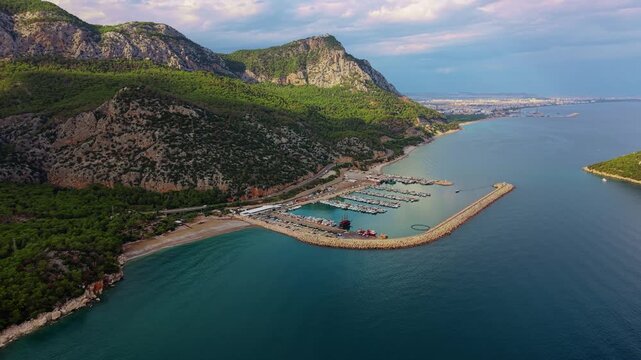 Fishing Shelter Poyraz Paşa Yat&ccedil;ılık in Konyaaltı, Antalya, Turkey, showing a curved harbor, forested mountains and calm Mediterranean water, cinematic aerial view