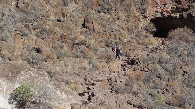 Alone, a woman hiker makes her way down a narrow stone trail etched into the side of a rugged mountain, Ansite fortress. The sun shines brightly, creating the perfect setting for an outdoor exploratio