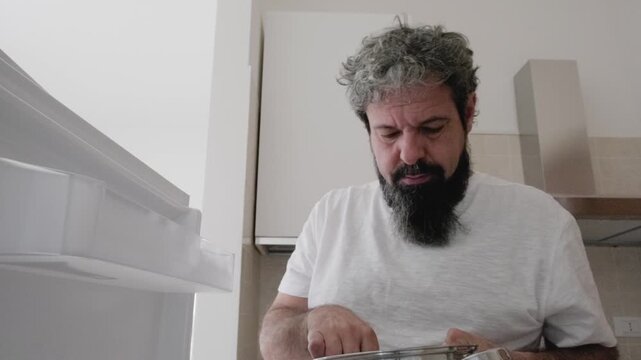 Bearded man opens refrigerator from interior POV, grabs cherries and eats them. Unique inside-fridge perspective captures snacking routine. Domestic kitchen lifestyle with fresh fruit.